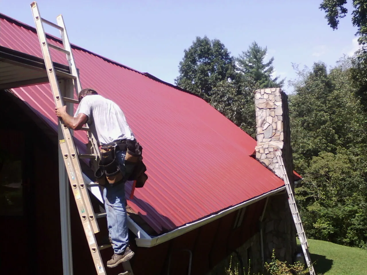 Roofer on a ladder beside a red metal roof and stone chimney — Soffit & Fascia Repair by Roofing Brothers in Port Orange